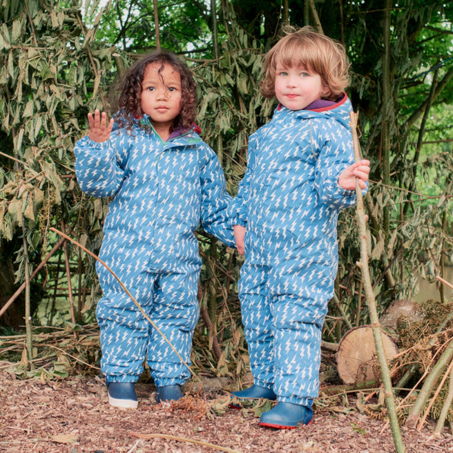 Two children wearing EcoWarm Waterproof Snowsuit Blue, standing in a woodland area, holding hands in their wellington boots.